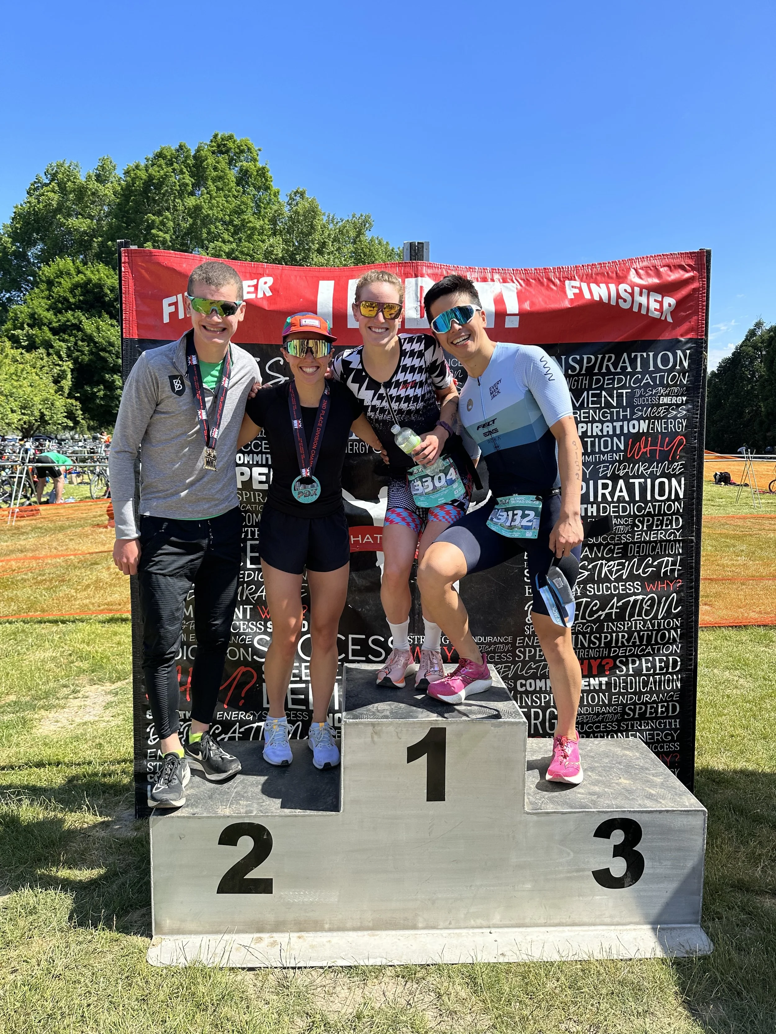 Group of four marathon runners standing on the winner's podium, celebrating their race, with a backdrop reading 'FINISHER' and a blue sky above.