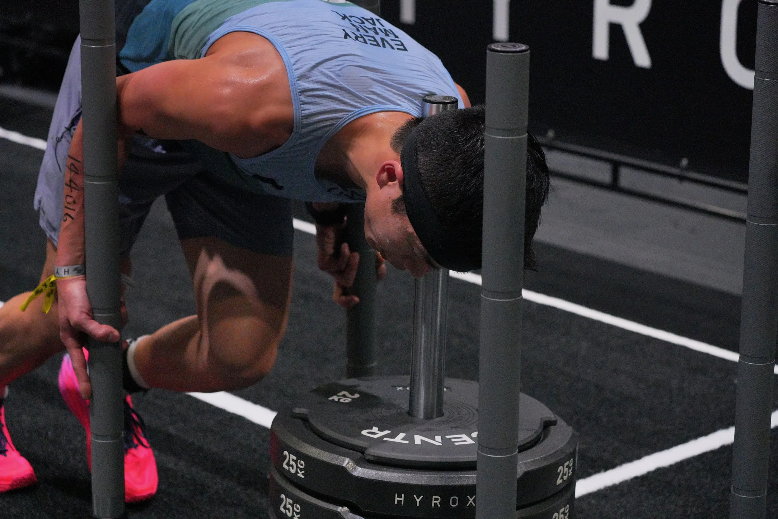 A man is performing a push-up exercise on a black gym floor with white lines, using a specialized weight vest and a barbell calorie wheel for support.
