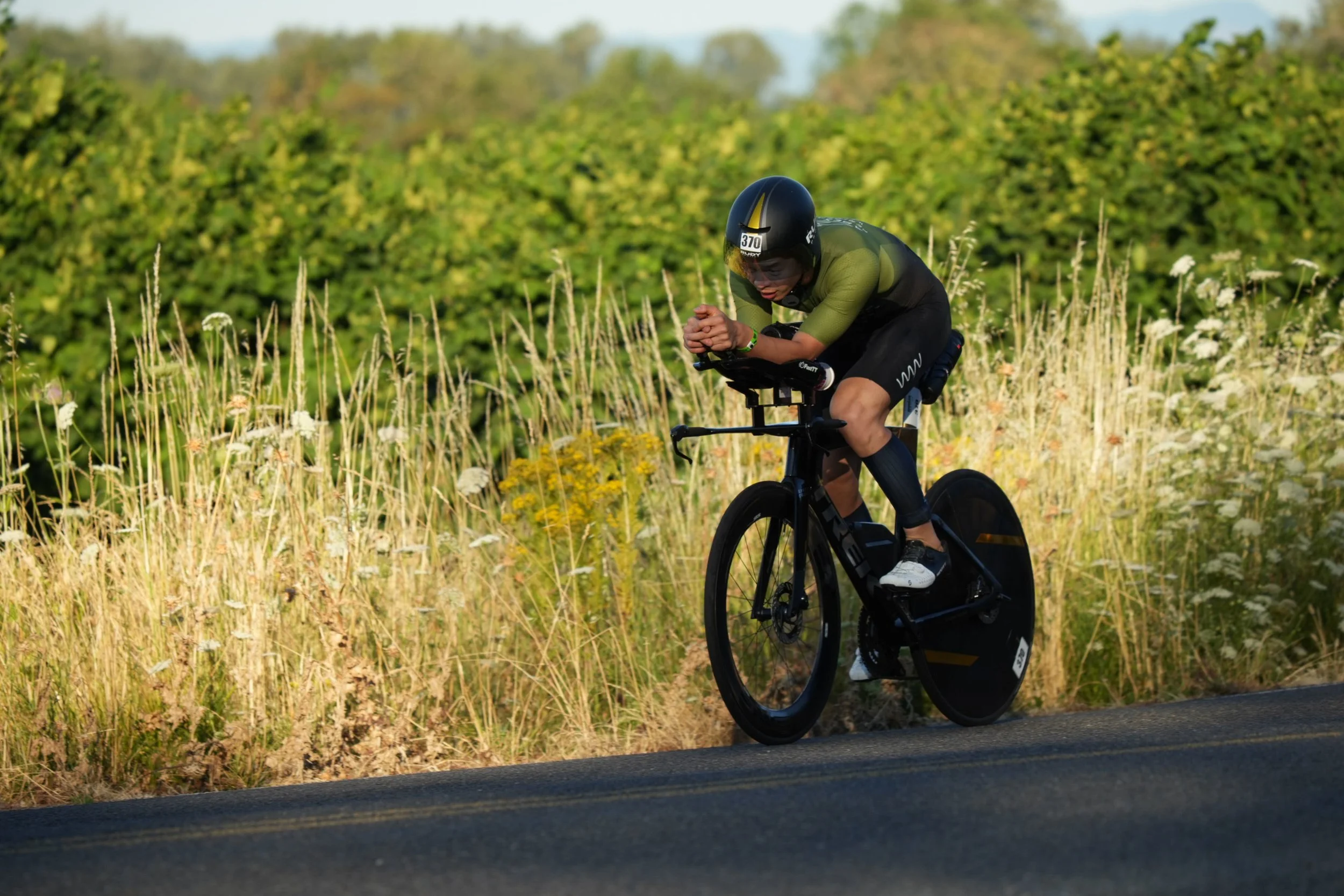 A cyclist in a green and black uniform riding a black time trial bike on a paved road through a field of tall grass and wildflowers, with green trees in the background.
