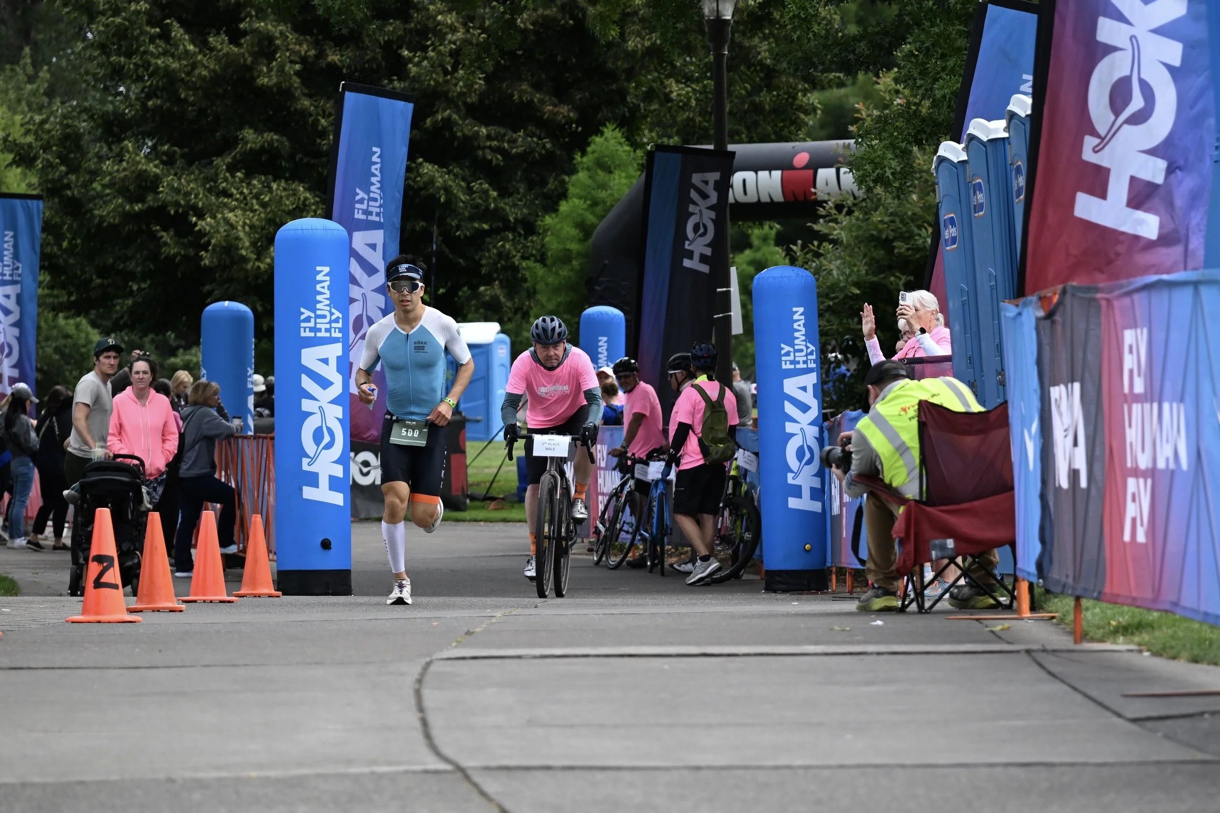 Participants finishing a race Oregon 70.3 IRONMAN event, with a cyclist running and others walking near the finish line, surrounded by blue banners and spectators.