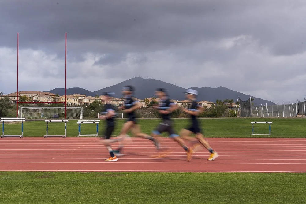 Four runners wearing black and blue athletic clothing and hats running on a red track field outdoors under cloudy sky, with hurdles, goalpost, green grass, and mountains in the background.