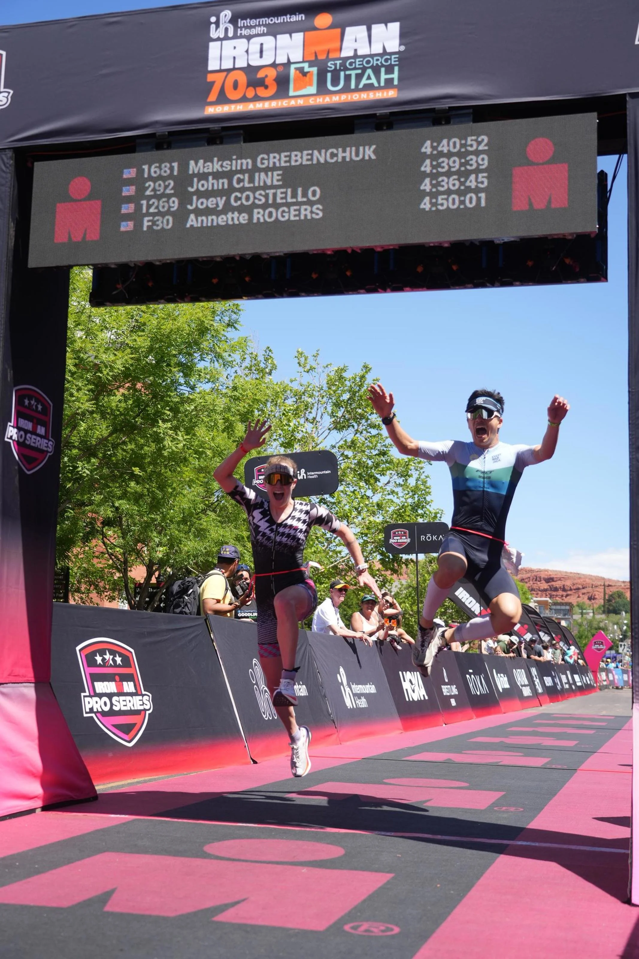 Two athletes crossing the finish line at the Ironman 70.3 triathlon in St. George, Utah, celebrating their achievement with smiles and raised arms. The digital clock above shows their finishing times, and spectators are visible in the background under a clear blue sky.