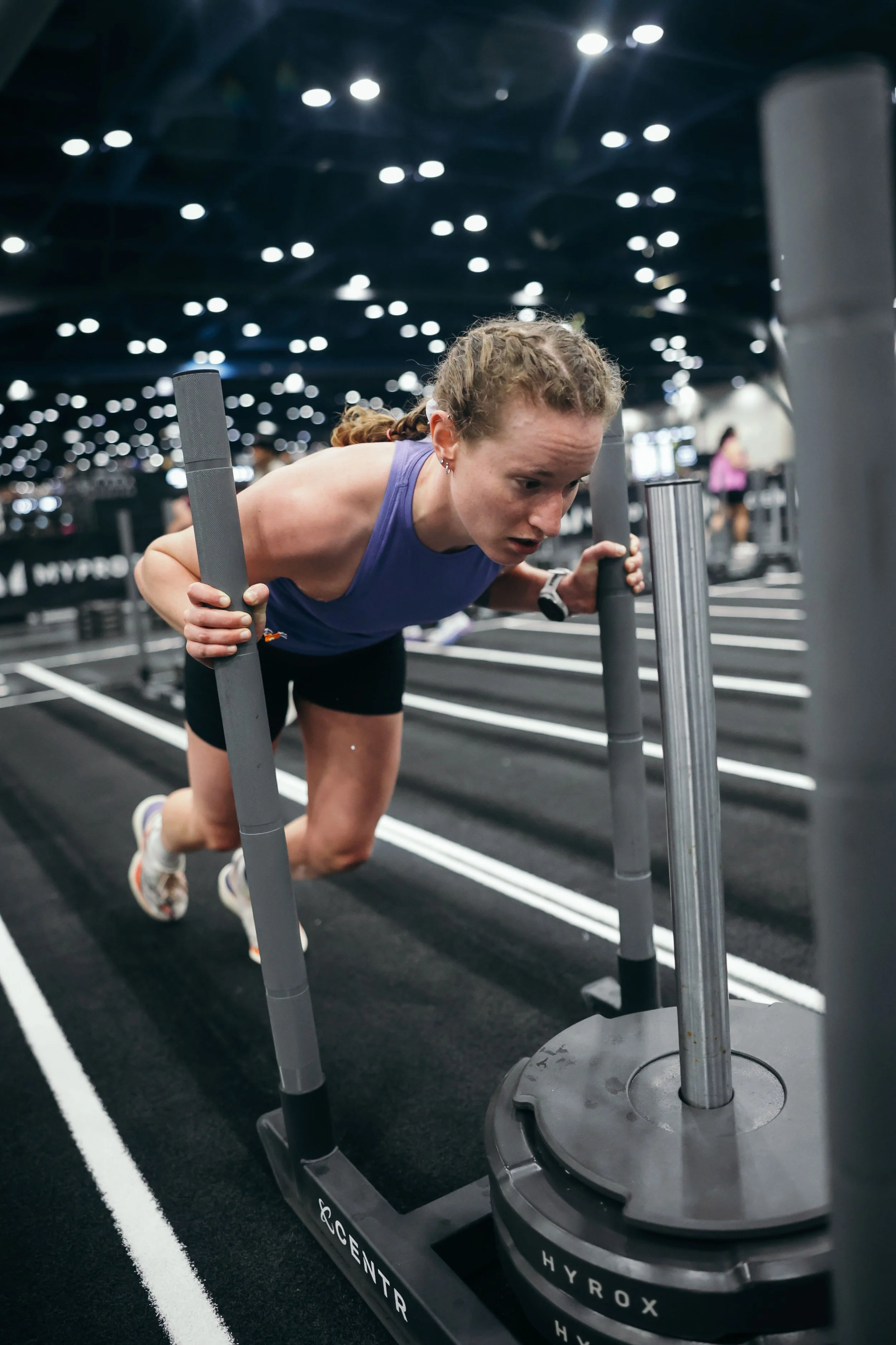 A woman in athletic clothing pushes a weighted sled on a track in a gym, with gym equipment in the background and ceiling lights above. Hyrox sled push