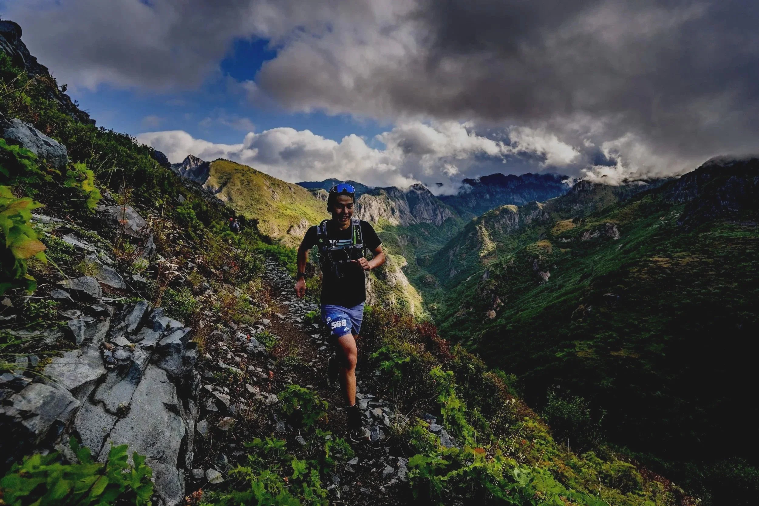 A man trail running on a narrow rocky trail in a lush green mountain landscape under a cloudy sky.