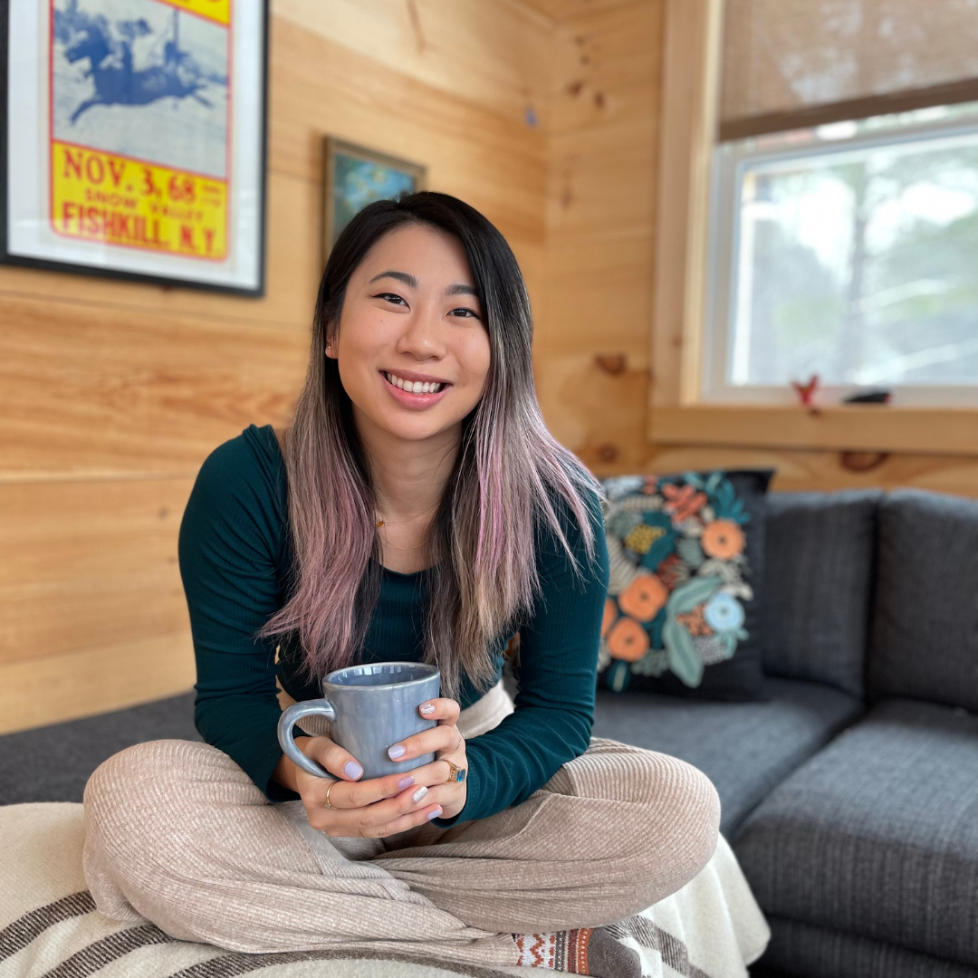 A young woman with long, wavy hair and a smile, sitting cross-legged on a cozy sofa holding a gray mug. The room has wooden walls, a window letting in natural light, a colorful floral pillow, and framed posters including a map in the background.