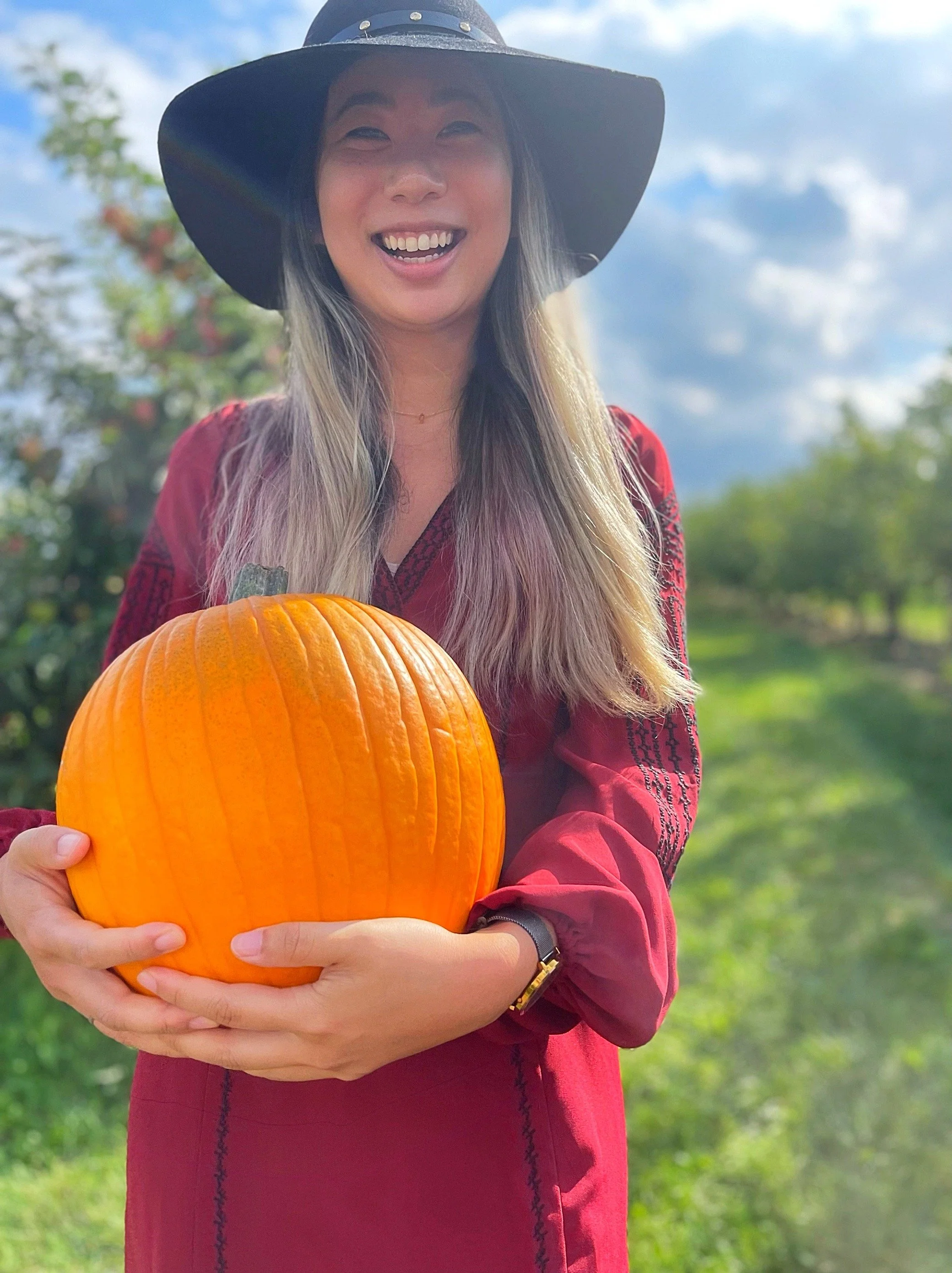 A woman with long hair wearing a wide-brimmed black hat and a red dress, smiling and holding a pumpkin outdoors with trees and a blue sky in the background.