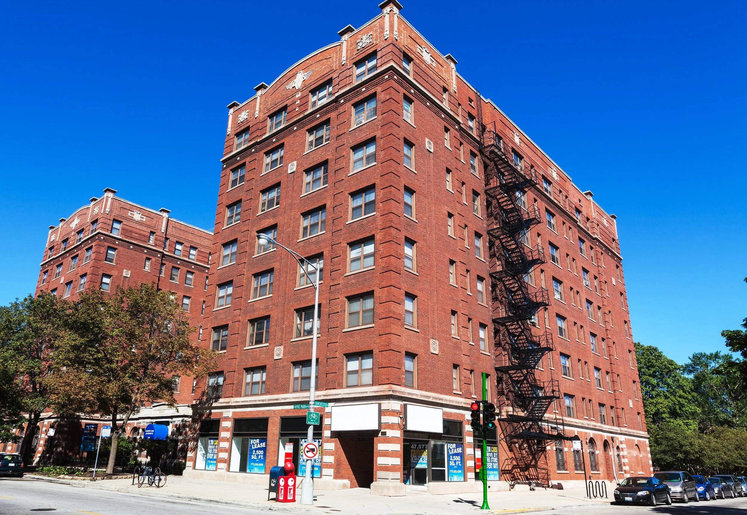 A large red brick residential apartment building with a fire escape on the side, surrounded by trees, street signs, and parked cars on a sunny day.