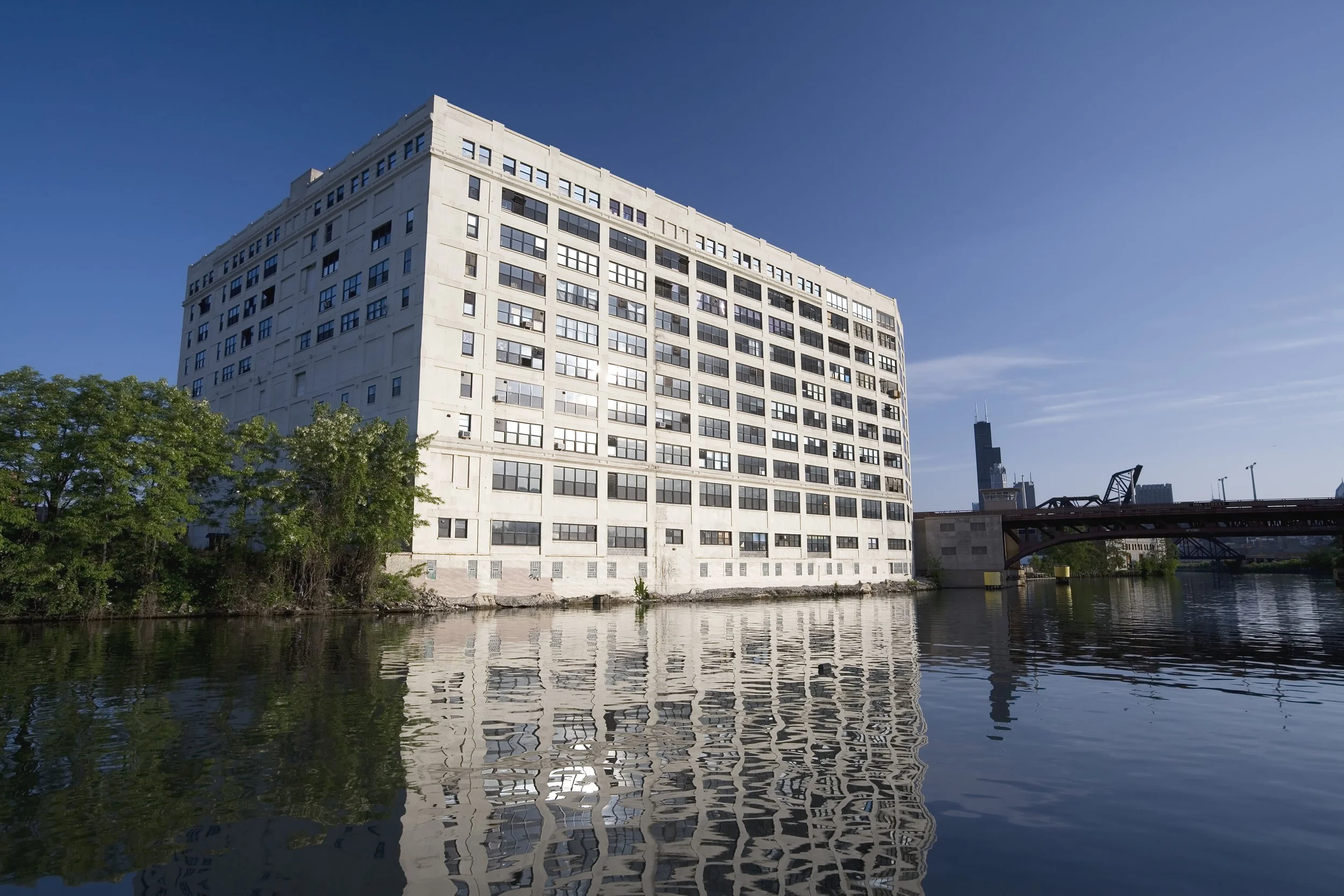 A large white multi-story building with many windows situated next to a body of water, with trees on the left, a bridge on the right, and a city skyline with a tall skyscraper in the background under a clear blue sky.