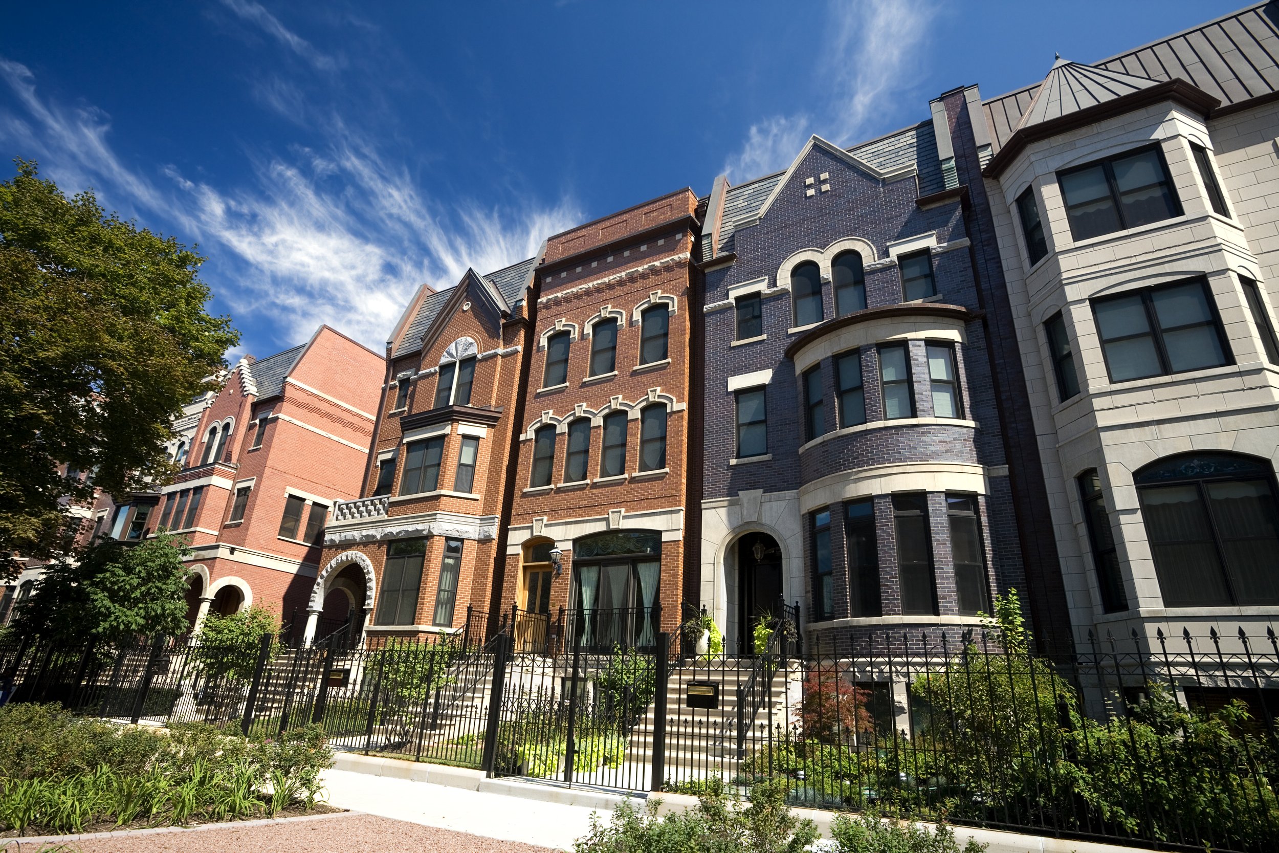 Row of Victorian-style townhouses with brick facades, arched windows, and decorative trim, enclosed by a black iron fence, under a bright blue sky with wispy clouds.