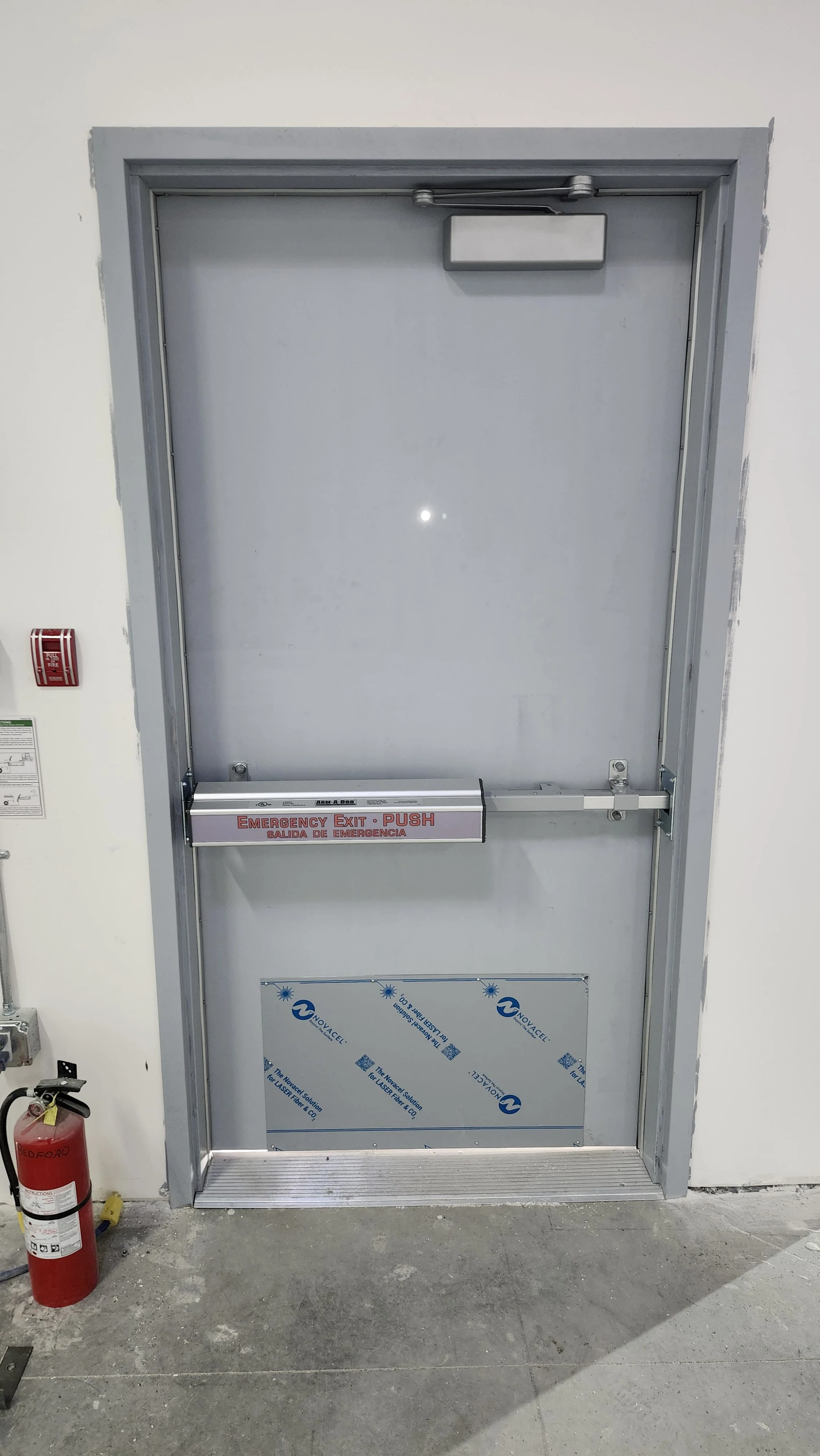 Gray metal door with an emergency push bar and a sign that reads 'Emergency Exit Push' and a fire extinguisher to the left of the door.