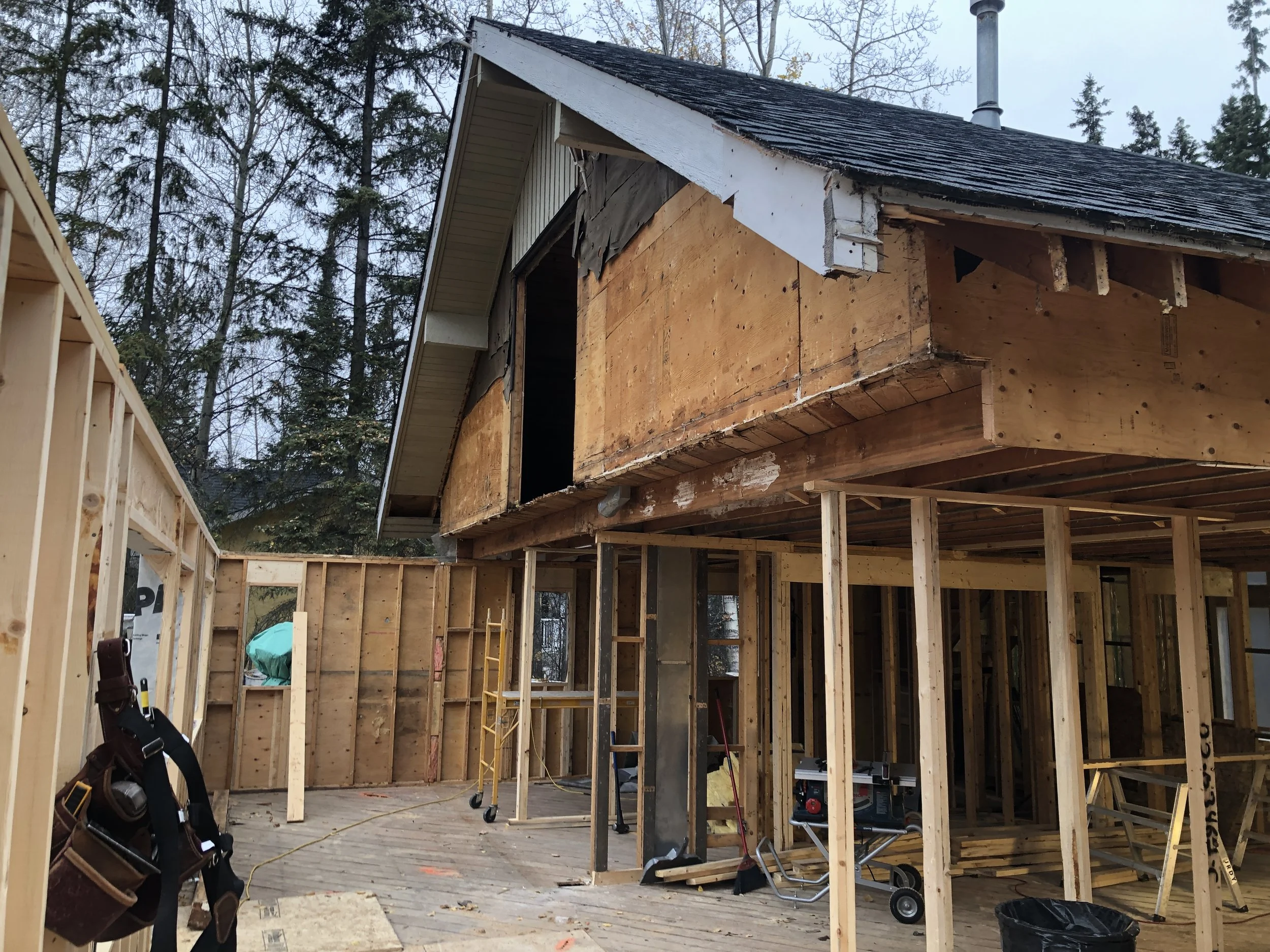 Construction site of a house with exposed wooden framing and partial exterior walls, in a wooded area with trees in the background.