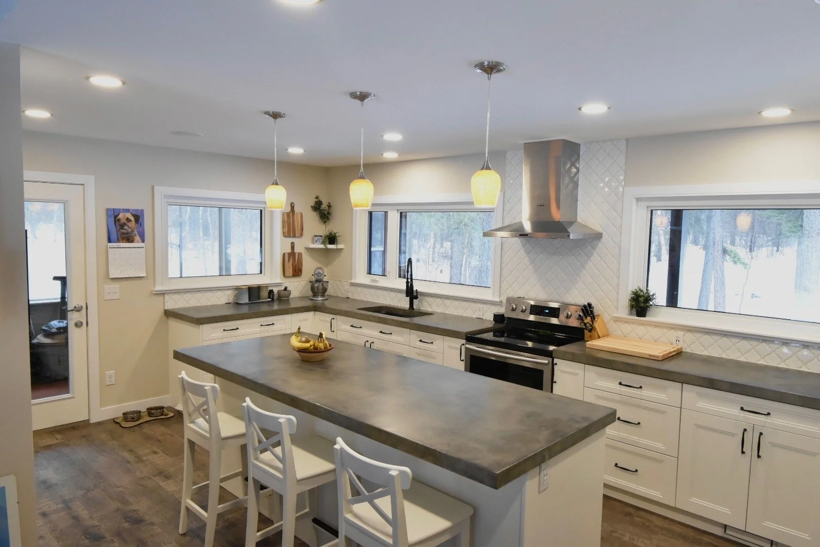 Modern kitchen with white cabinets, gray countertops, and stainless steel appliances. Three pendant lights hang above a central island with three white chairs. Large windows and a door reveal a snowy outdoor scene.