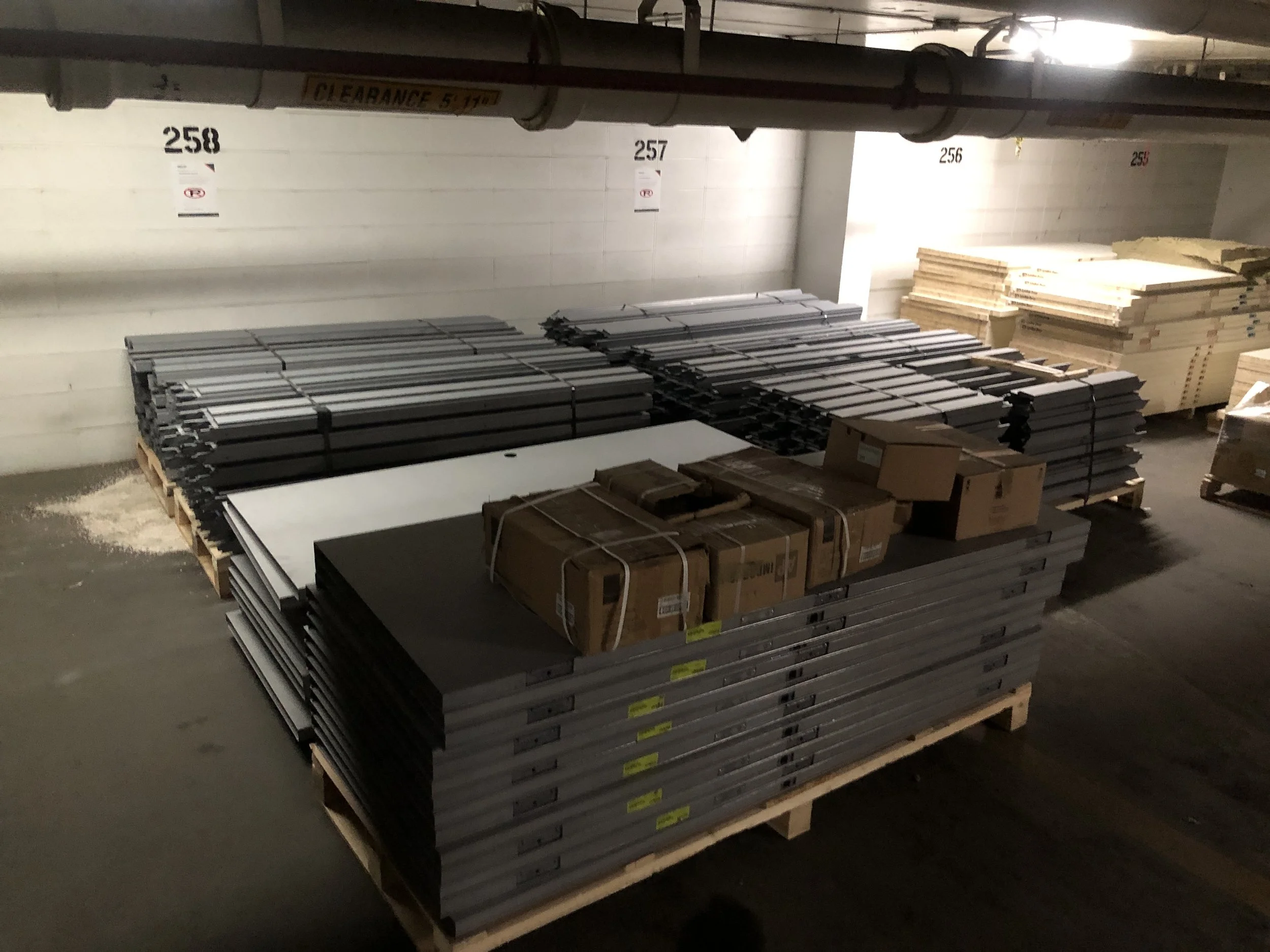 Pallets of stacked black, white, and wooden materials stored in an underground parking garage with numbered sections and pipes overhead.