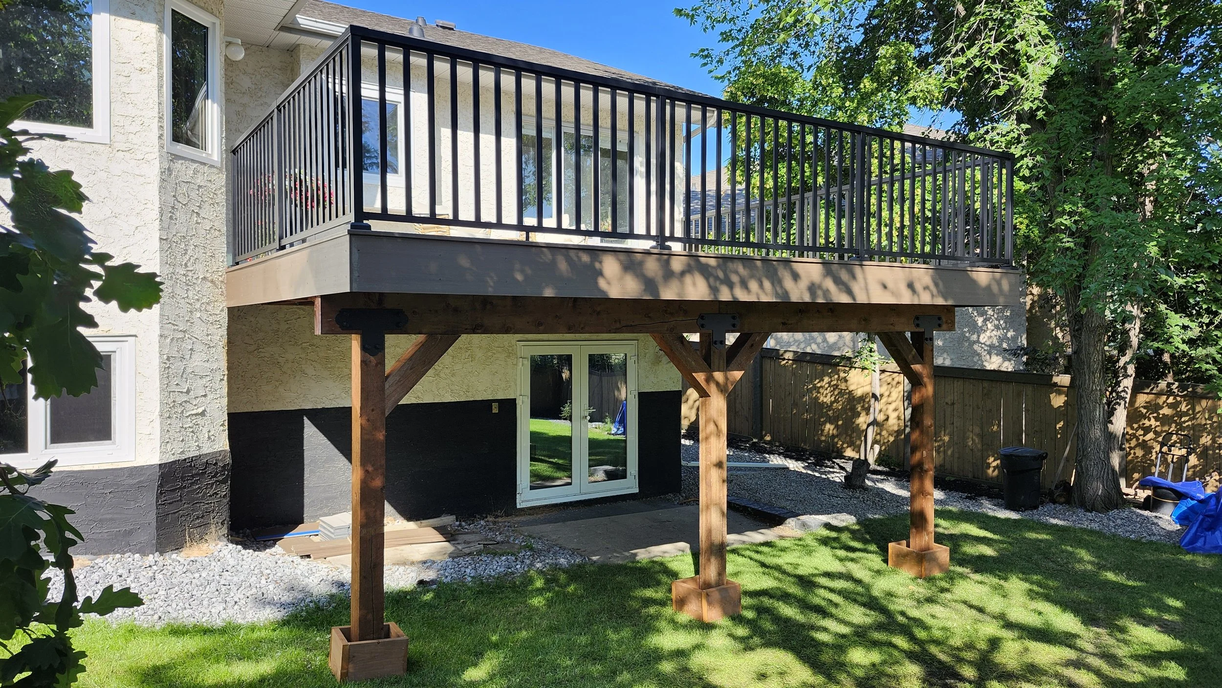 A new wooden deck with black metal railing built above a lower level of a house's backyard, supported by large wooden posts. The house has white textured walls with black paint at the bottom, and there are trees and a fenced yard with grass and gravel.