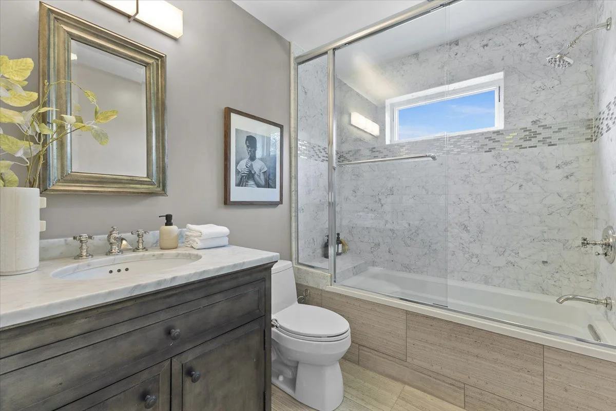 Modern bathroom with a marble bathtub, glass shower doors, a vanity with a marble countertop, a framed mirror, a black and white portrait photograph on the wall, and a window with blue sky outside.