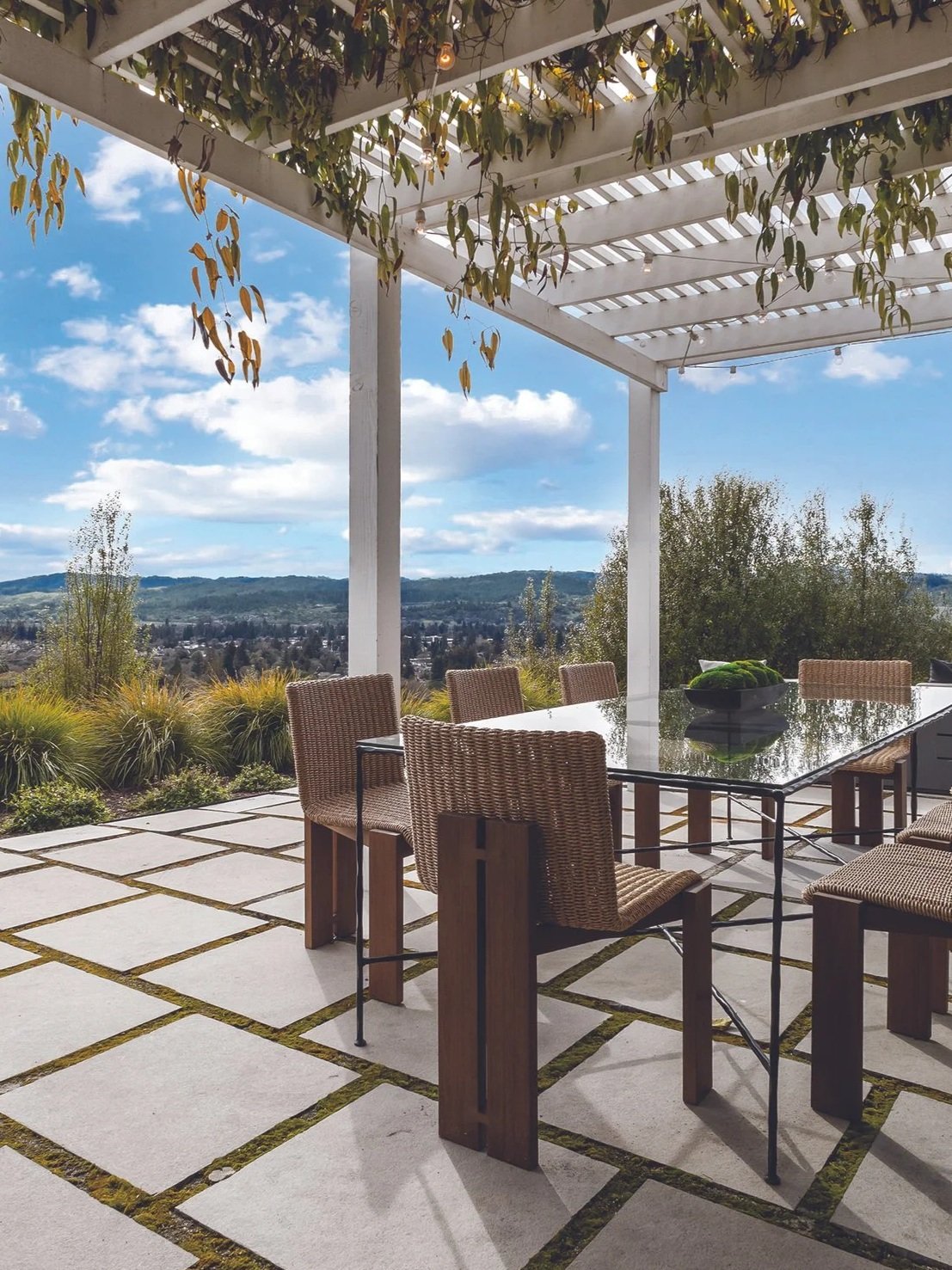 An outdoor patio with a large glass-topped table surrounded by wicker chairs, overlooking a scenic view of rolling hills and a blue sky with clouds. A white pergola with hanging leaves provides shade.