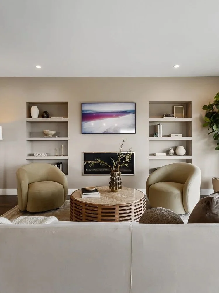 Living room with two beige armchairs, a round wooden coffee table with a vase and books, a white sofa in the foreground, a wall-mounted fireplace, a flat-screen TV above, built-in shelves holding decorative items, and a small plant on the right.