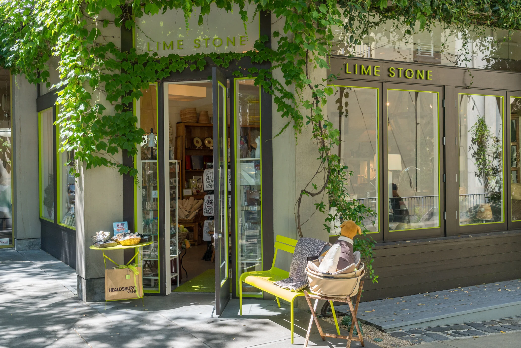 Exterior of a boutique store named Lime Stone with large glass windows and an open door, decorated with green ivy. Outside, there are a yellow bench, a small table with a basket of items, and a pet dog in a stroller.