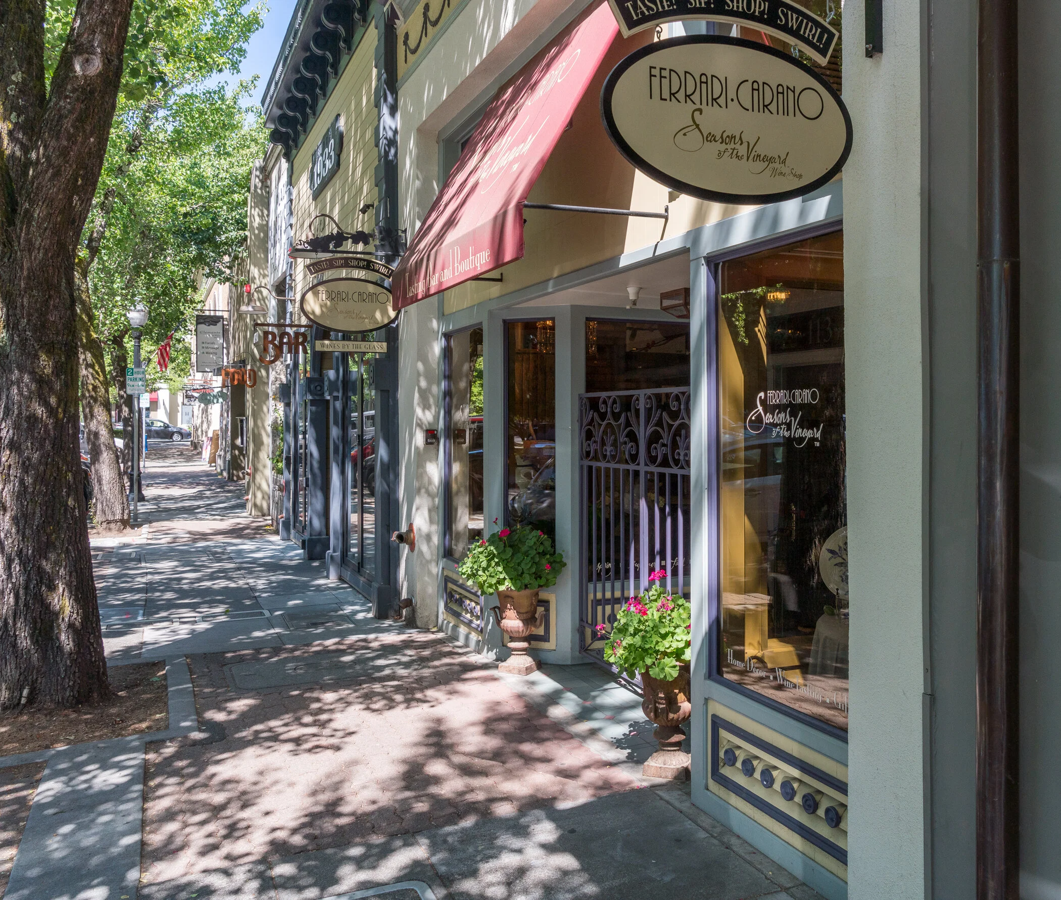 A sunny street view with storefronts and trees, featuring a winery shop with potted plants and a red awning.