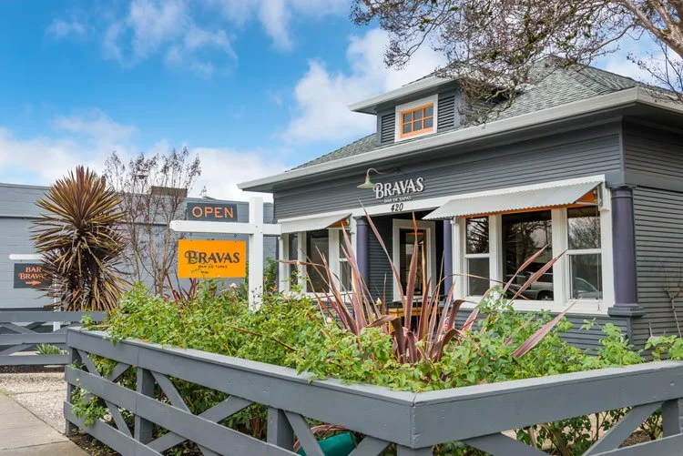 Blue building with white trim, labeled 'Bravas' at 420, featuring a front porch, surrounded by greenery and an overcast sky.