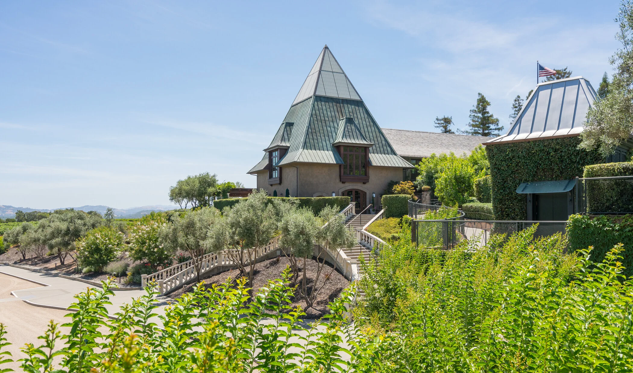 A house with a steep, pointed, metal roof, situated on a landscaped hill with stairs, surrounded by greenery and trees, with a clear blue sky in the background.