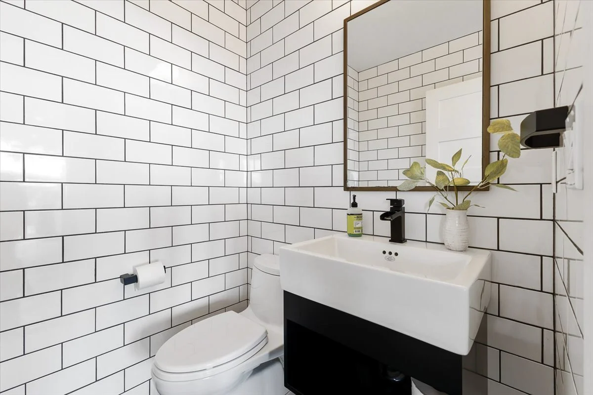Modern bathroom with white subway tile walls, a wall-mounted sink with a black faucet, a mirror, a decorative vase with green leaves, a toilet, a toilet paper holder, and a wall-mounted black tissue box.