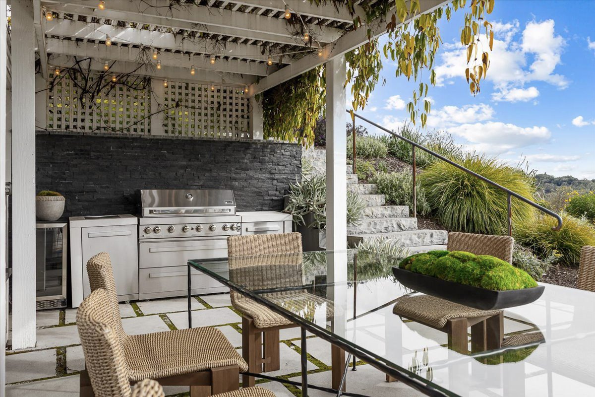 Outdoor patio area with a glass dining table, wicker chairs, outdoor kitchen with a grill, and stairs leading to a garden with plants and a blue sky with clouds.