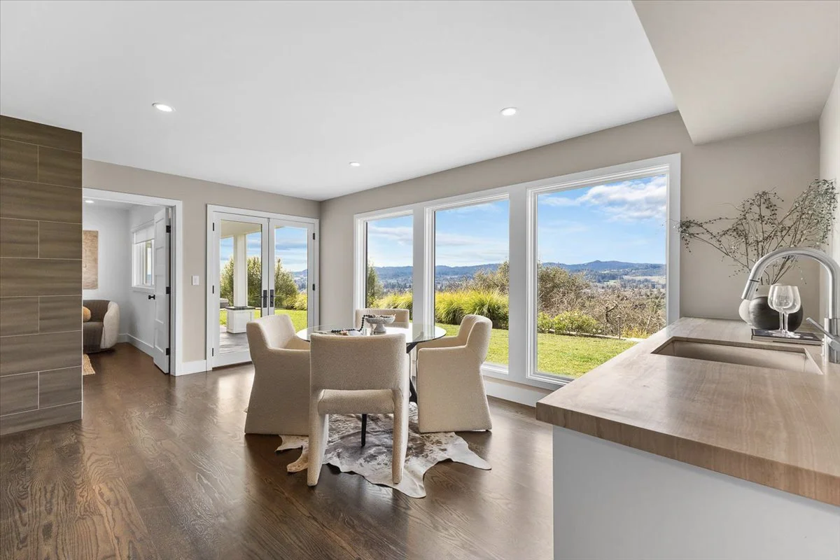 Bright dining area with large windows overlooking a scenic landscape, featuring beige chairs around a glass table and a natural wood countertop with a sink.