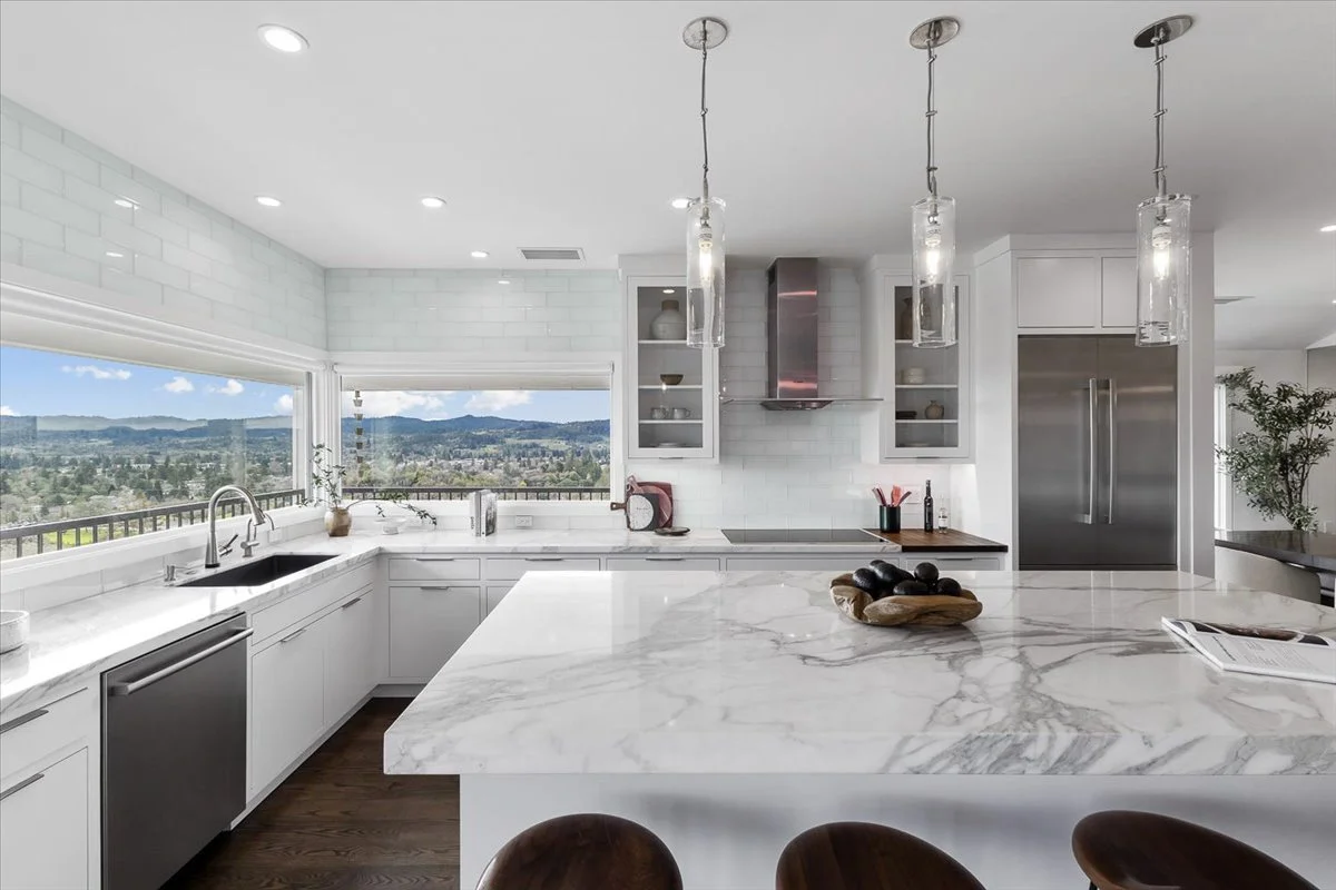 Modern kitchen with white cabinets, marble countertops, stainless steel appliances, and large windows showing a distant landscape.