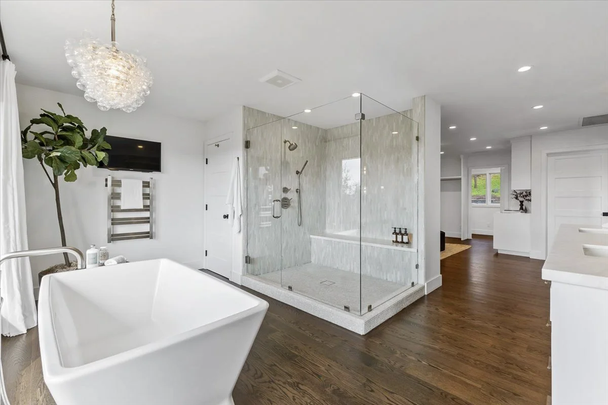 Modern bathroom with a large, white freestanding bathtub, a glass-enclosed shower with marble tiles, and wooden flooring. There is a small flat-screen TV on the wall, a green houseplant, and a lighting fixture hanging from the ceiling.