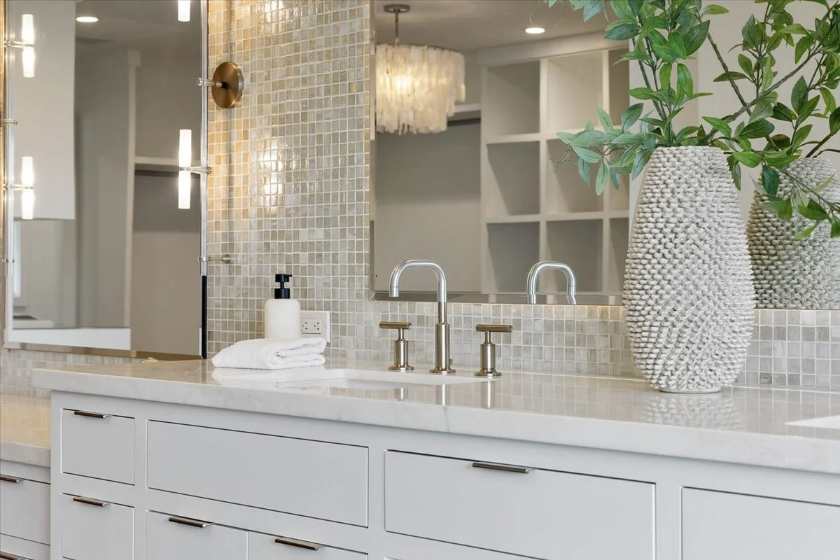 A modern bathroom vanity with a white marble countertop, two gold faucets, and a large mirror. There are two large textured vases with green leaves, a soap dispenser, and a towel on the countertop. The background features a tiled wall and a ceiling l