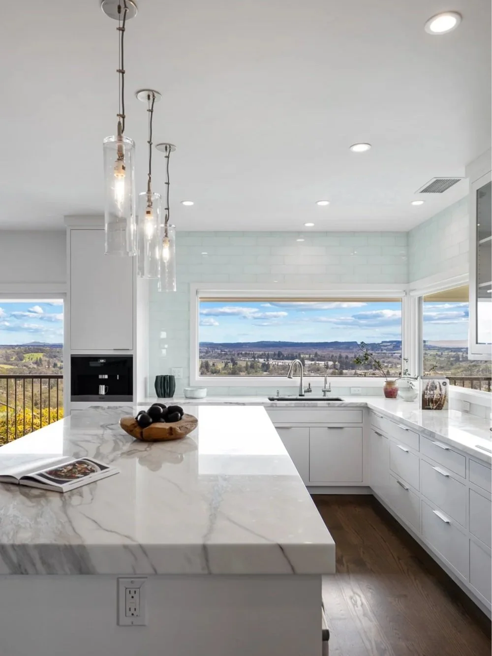 White modern kitchen with large marble island, view of landscape through windows, pendant lights over island, and organized countertops.