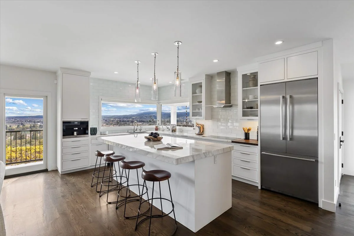 Modern kitchen with white cabinets, a marble island, stainless steel refrigerator, and large windows showing a scenic outdoor view.