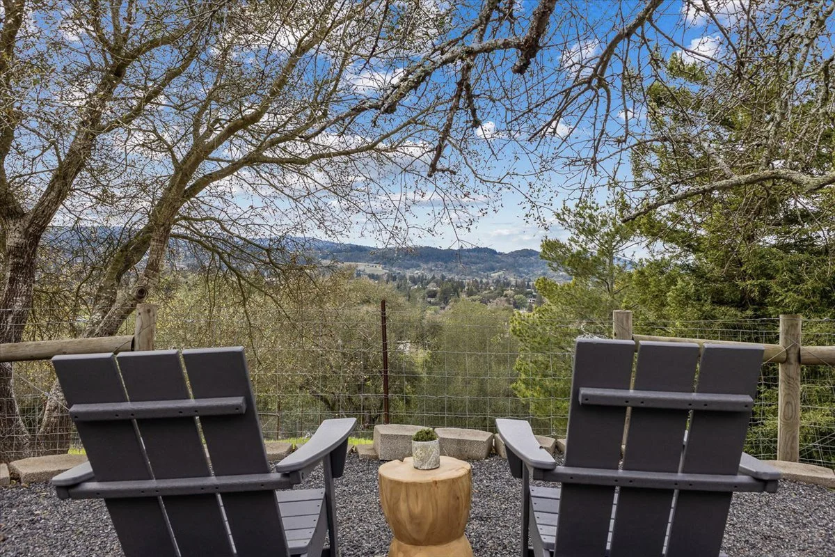 Two black Adirondack chairs facing a scenic landscape with rolling hills, trees, and a cloudy sky, on a gravel patio with a small wooden side table between them.
