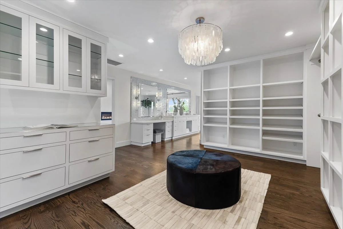 A spacious, modern living room with white built-in cabinets, a large open white bookshelf, a round dark-colored ottoman, a cream-colored rug, a chandelier, and a mirror reflecting the room's plants and windows.