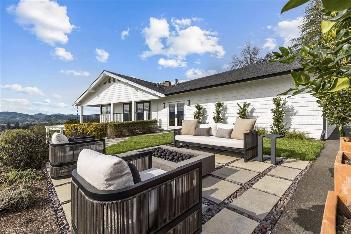 Outdoor patio with modern furniture, including a sofa, armchairs, and a fire pit, next to a white house with large windows and a small balcony. The patio is landscaped with grass, small bushes, and pavers, with a scenic view of hills and blue sky wit
