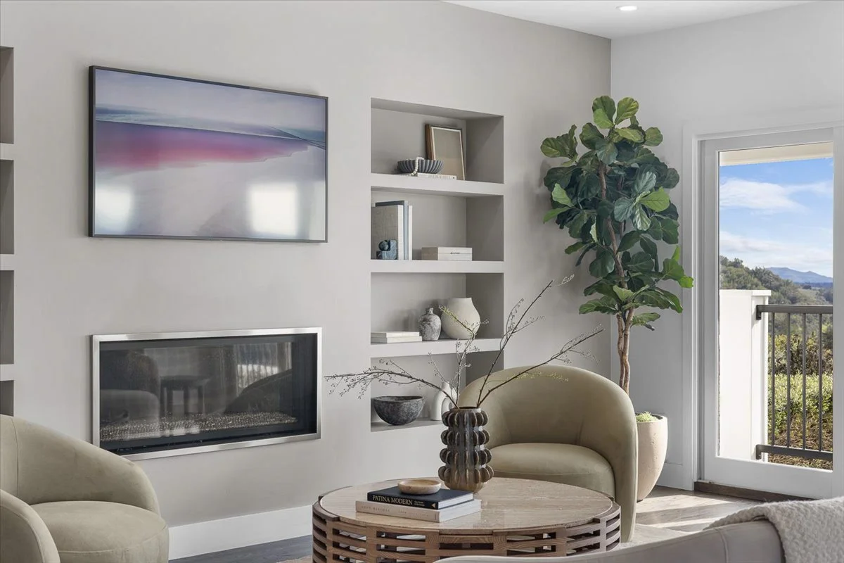 Living room with beige armchairs, a wooden coffee table with a brown vase and books, a built-in white shelf with decor, and a large potted plant near a sliding door with a view of a balcony and landscape outside.