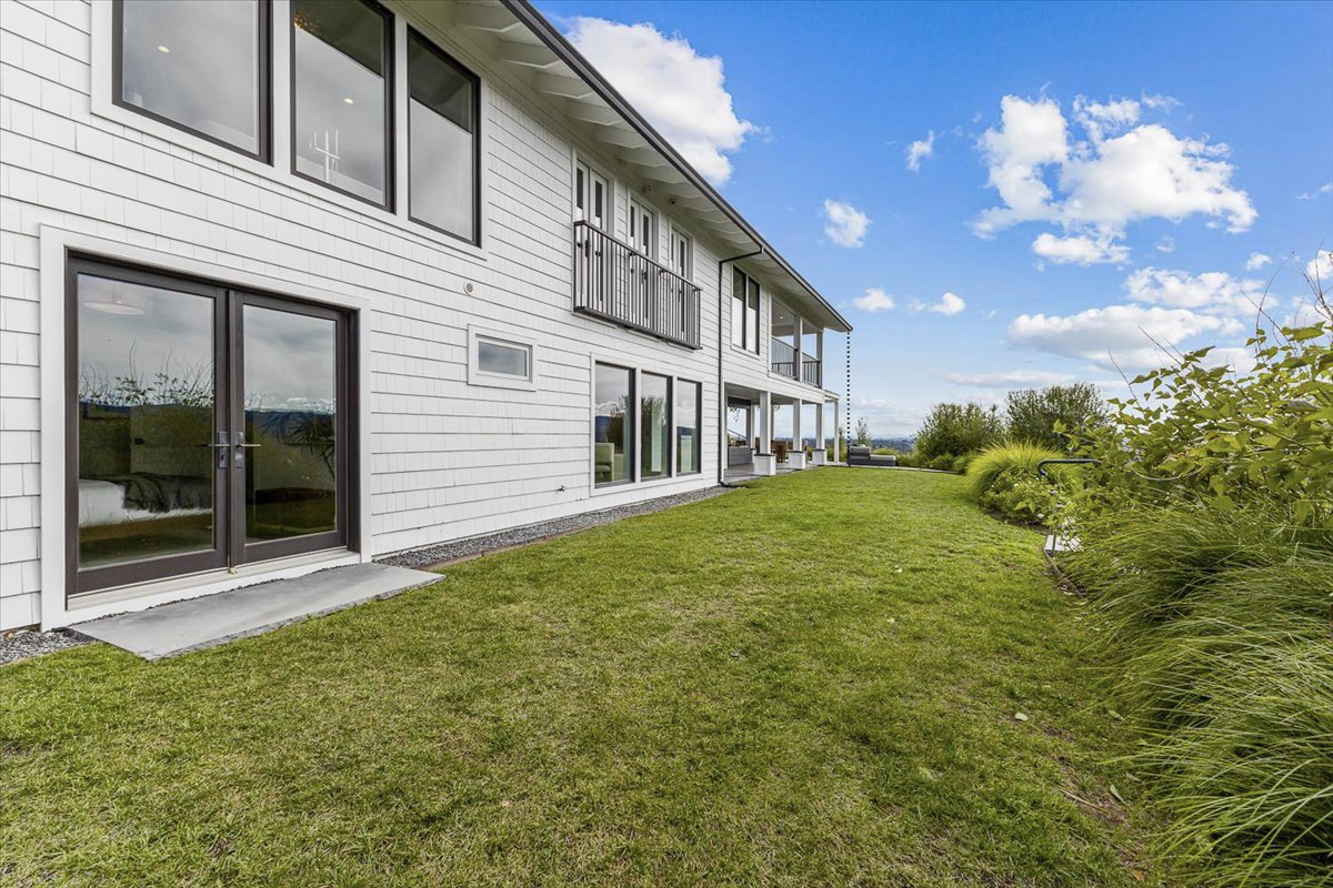 View of a modern two-story house with white siding, large windows, and a backyard with green grass and bushes, under a partly cloudy sky.