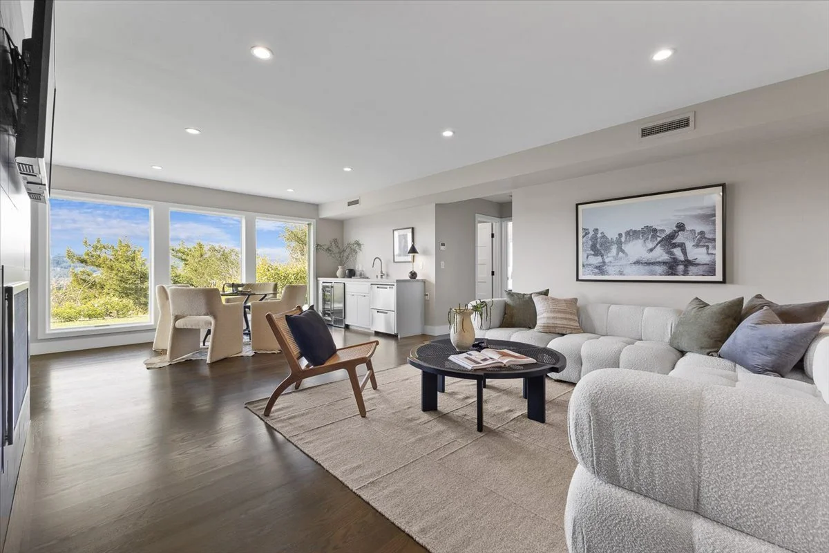 Bright living room with large windows showing greenery outside, white sectional sofa with throw pillows, wooden armchair, black coffee table with books, and a dining area with four chairs in the background.