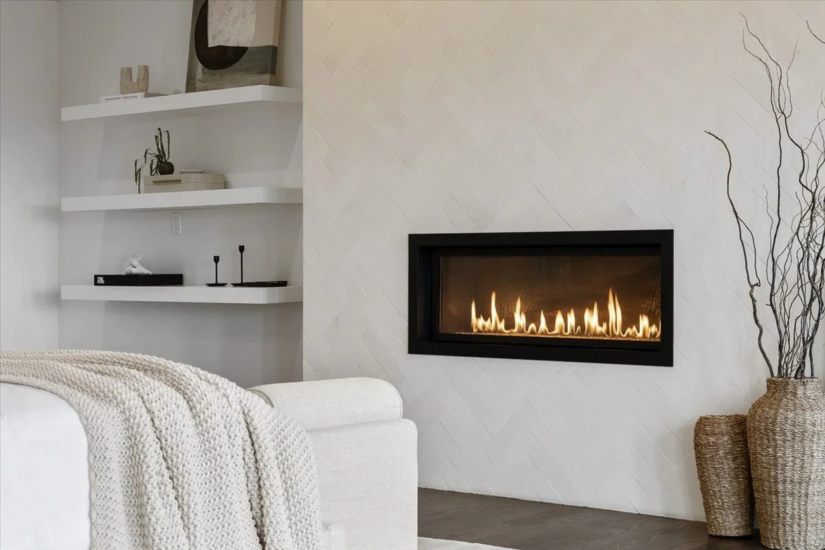 Modern living room with a white couch, a knitted throw blanket, a built-in fireplace with flames, white floating shelves with decorative items, and woven baskets with tall branches.
