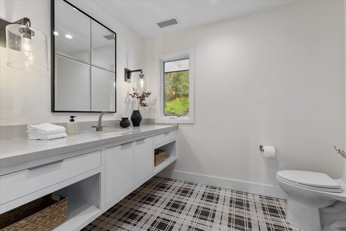 Modern bathroom with white vanity, black-framed mirror, black light fixtures, window showing green outdoors, and plaid floor.