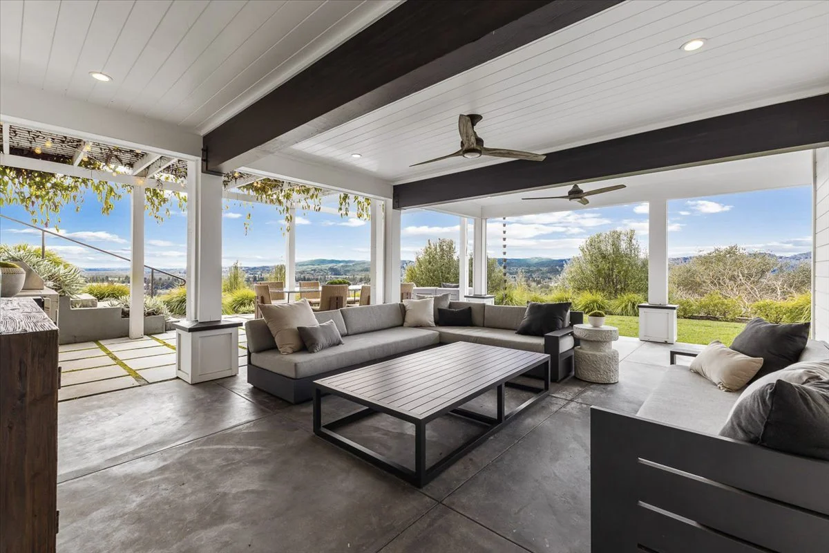 Covered patio with outdoor seating including a large L-shaped sofa and a square coffee table, overlooking a scenic landscape with trees and hills in the distance, and open sky with scattered clouds.