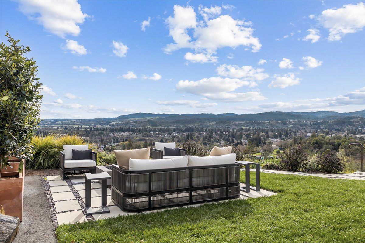 Outdoor patio with modern seating area, including a black textured couch with beige and black pillows, a matching armchair, and two small side tables, overlooking a scenic view of rolling hills and a partly cloudy sky.