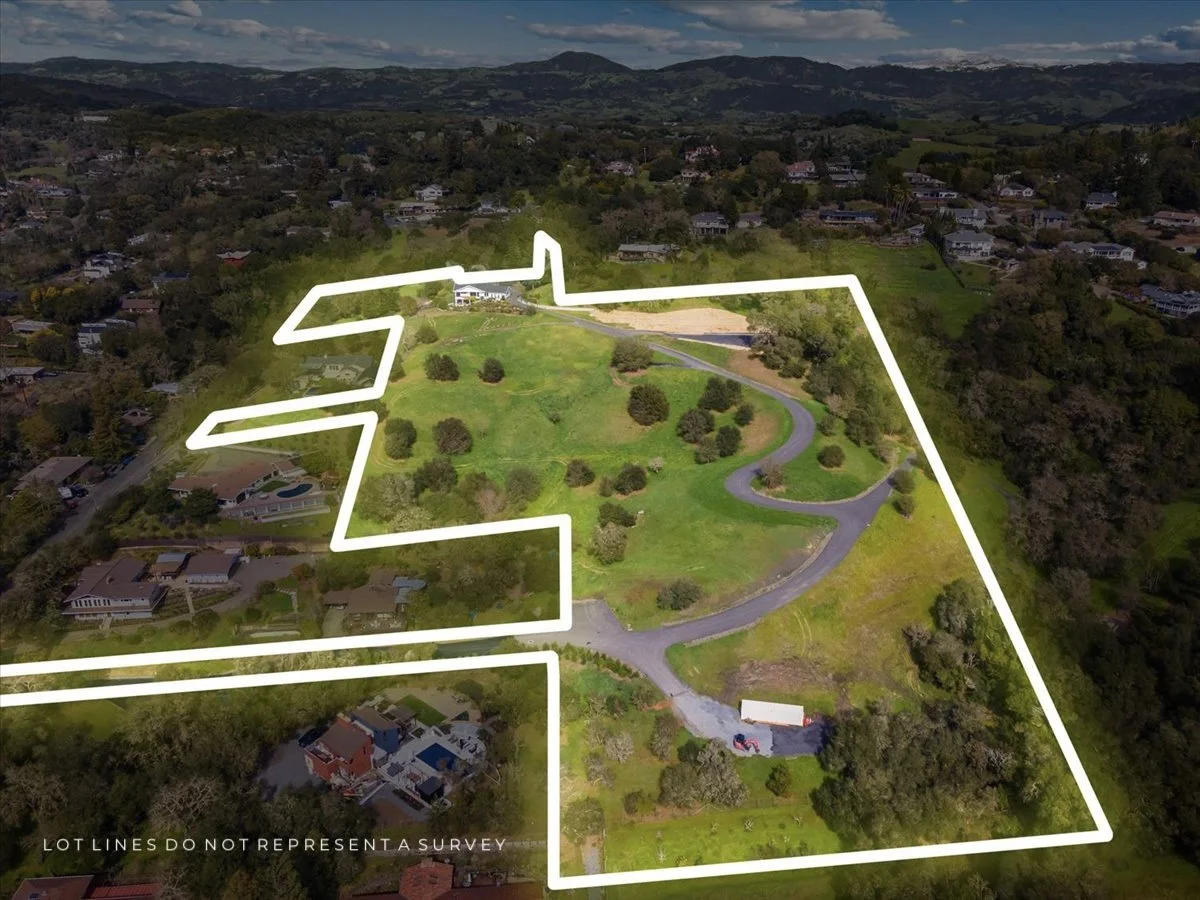 Aerial view of a large property outlined with white lines indicating boundaries, featuring a winding driveway, trees, grassy areas, and nearby houses in a hilly landscape.