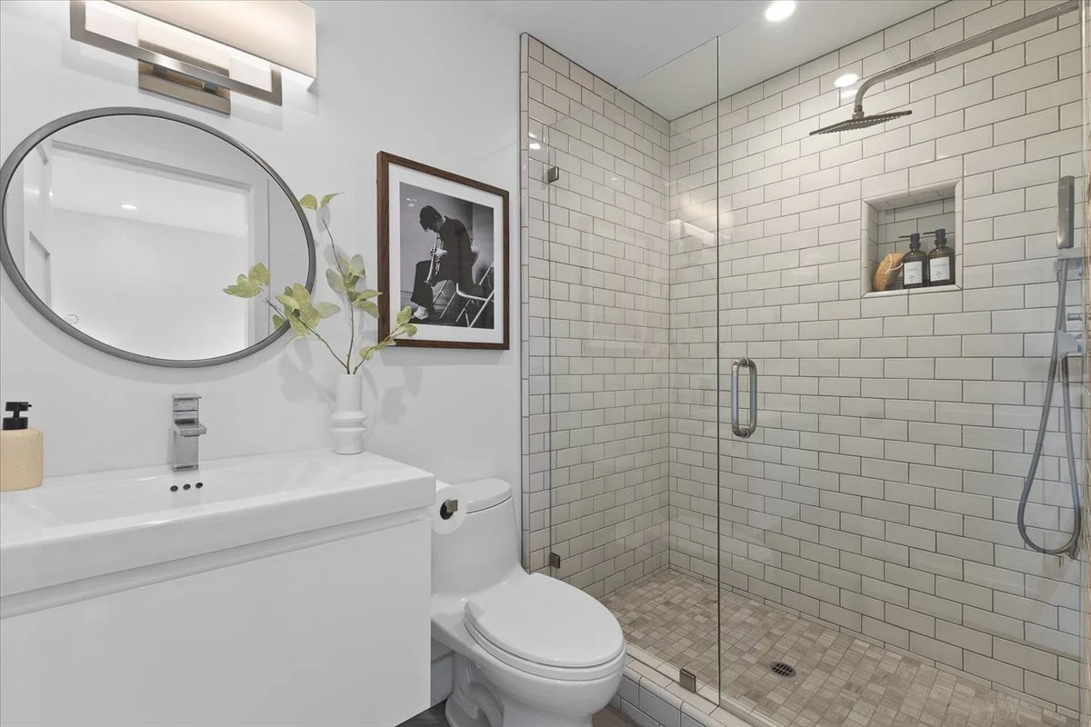Modern bathroom with white tile walk-in shower, toilet, and sink. Decor includes a round mirror, a framed black-and-white photo, and a vase with green leaves.