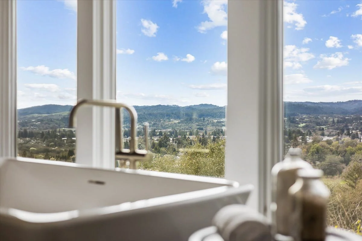 Kitchen sink with a view of a bright blue sky, scattered clouds, and distant mountains seen through large windows.