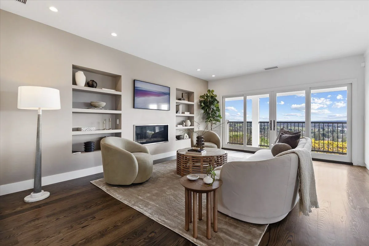 Living room with large sliding glass doors showing a scenic view of the outdoors, beige walls, a white ceiling with recessed lighting, a floor lamp, a white curved sofa with pillows, an armchair, a round coffee table, and built-in wall shelves with d