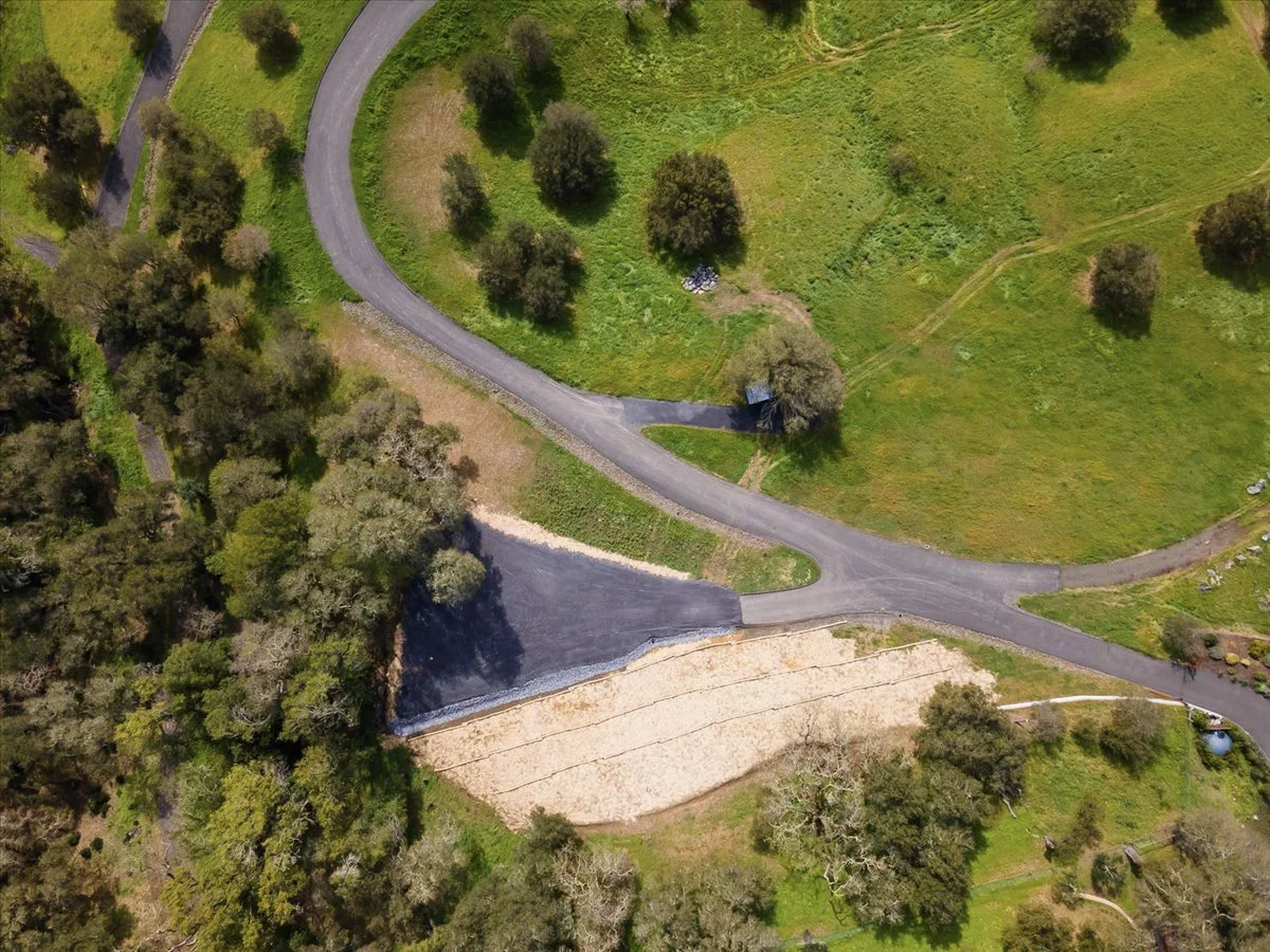 A winding paved road in a green landscape with trees, grass, and a small dirt area.