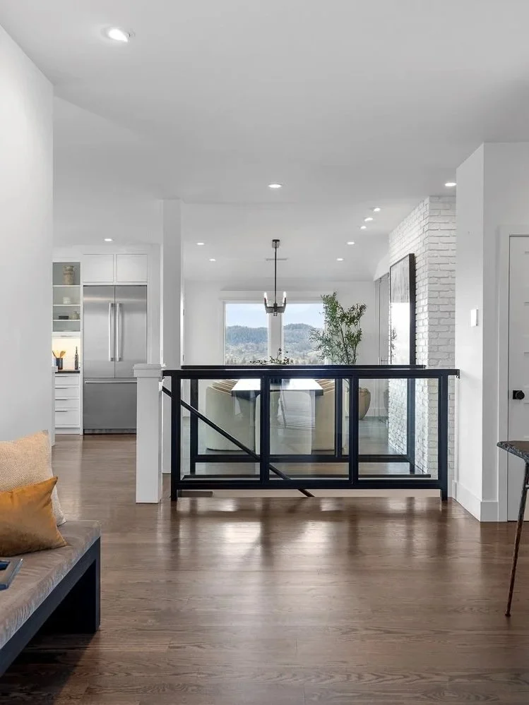 Living room with wood floor, white walls, kitchen area in the background, and a dining table with chairs behind a glass railing.