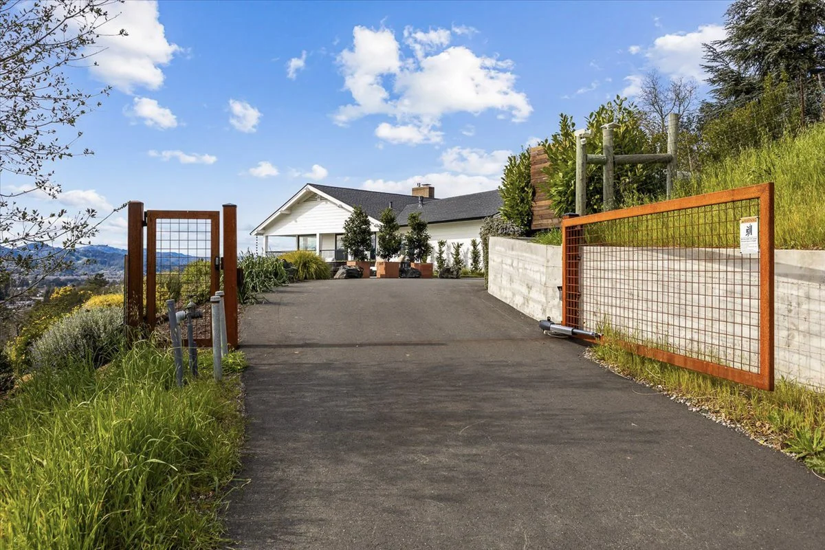 A paved driveway leading to a white house with a black roof, surrounded by green plants and trees, with a blue sky and white clouds overhead. There are orange gates on either side of the driveway.